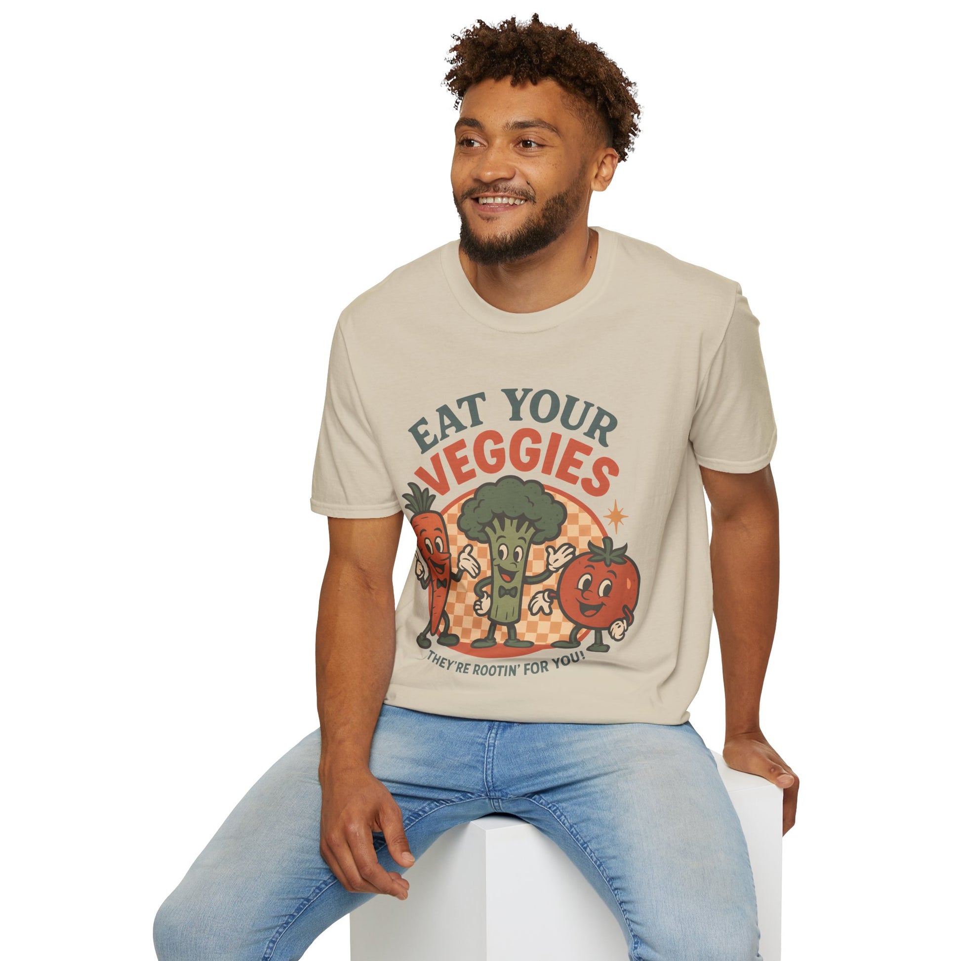 Male model wearing a cream “Eat Your Veggies” Vegan Vegetarian Veggie Vegetable Tshirt with smiling carrot, broccoli and tomato characters, seated on a white block.
