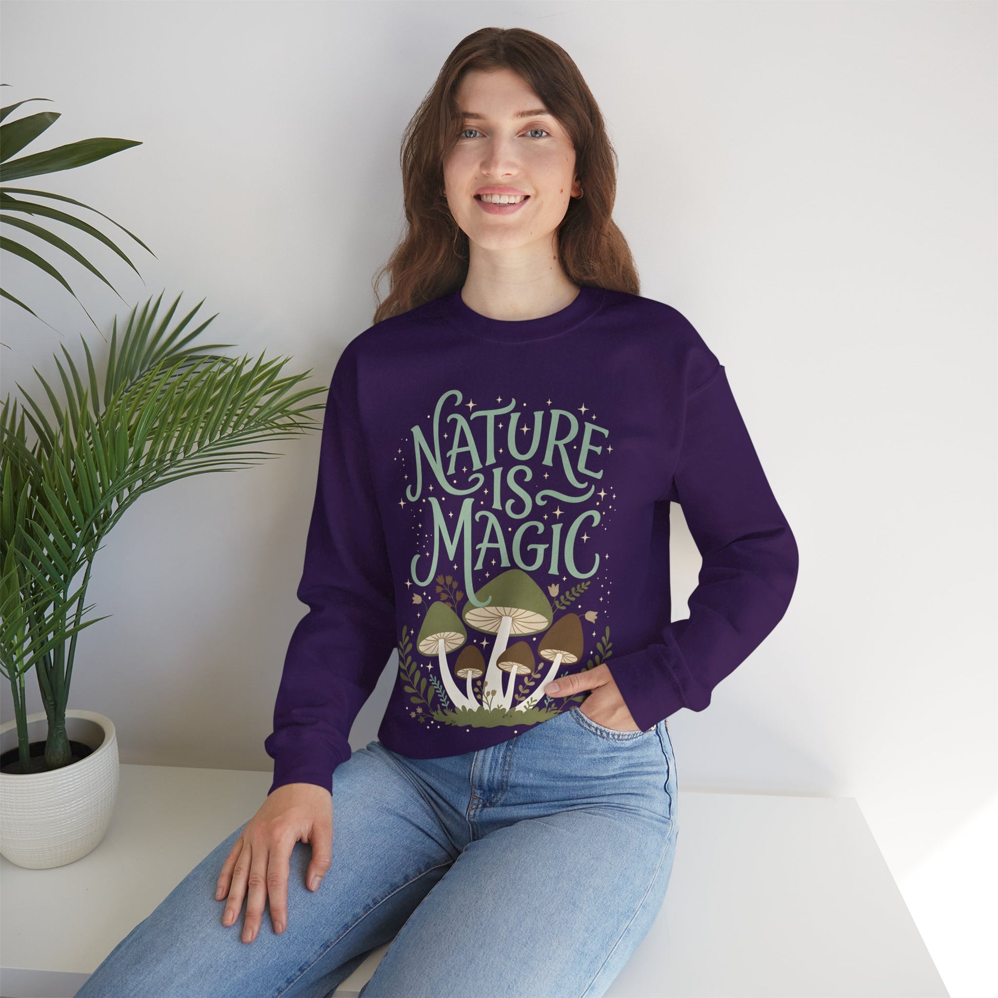 Smiling model sitting beside a potted plant in a black “Nature Is Magic” mushroom sweatshirt, featuring earthy forest mushrooms and leafy nature artwork on the front.