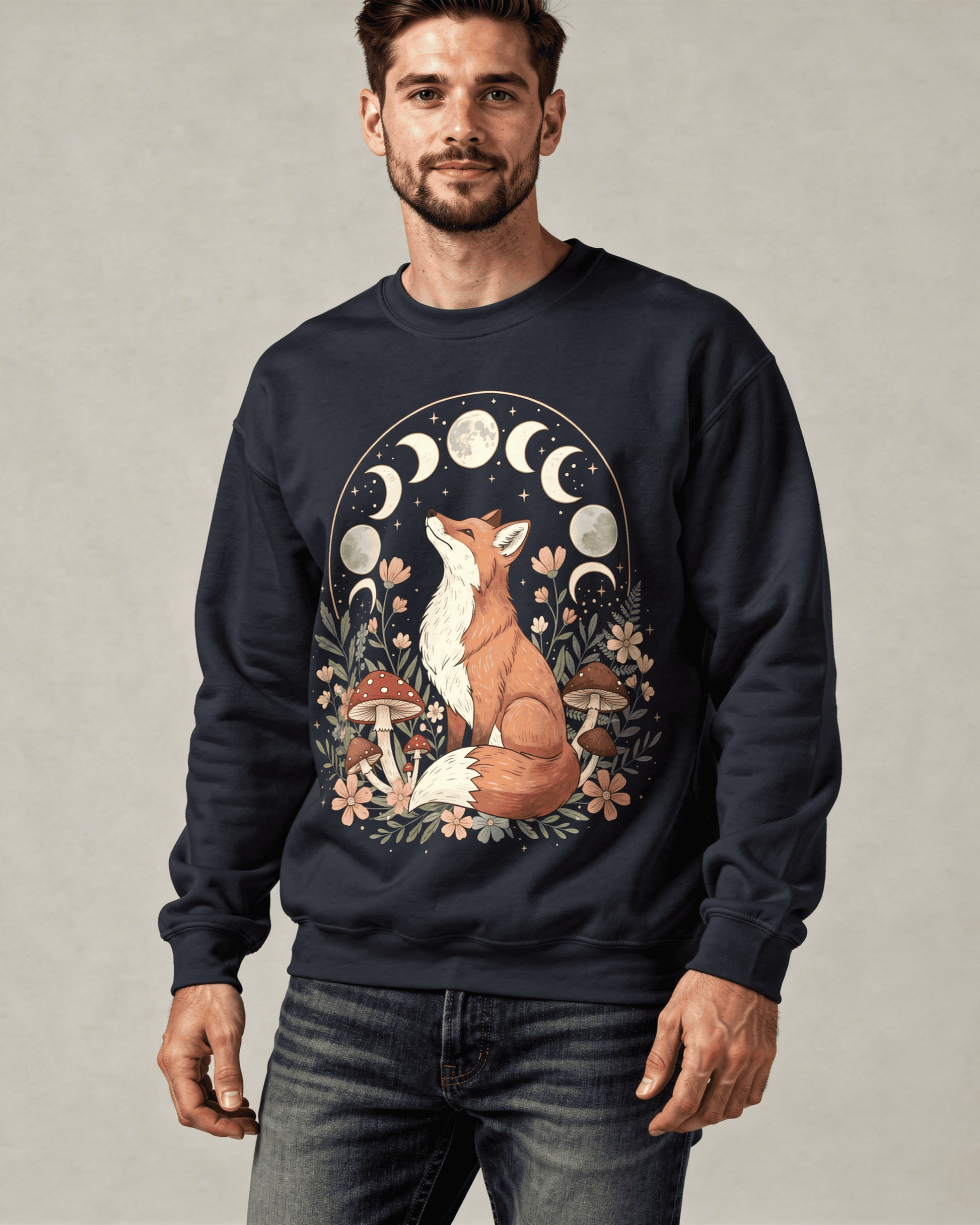 A man with a beard models a dark crewneck sweatshirt with a detailed cottagecore design. An orange fox is centered in a botanical scene, looking up at the complete moon phases cycle.