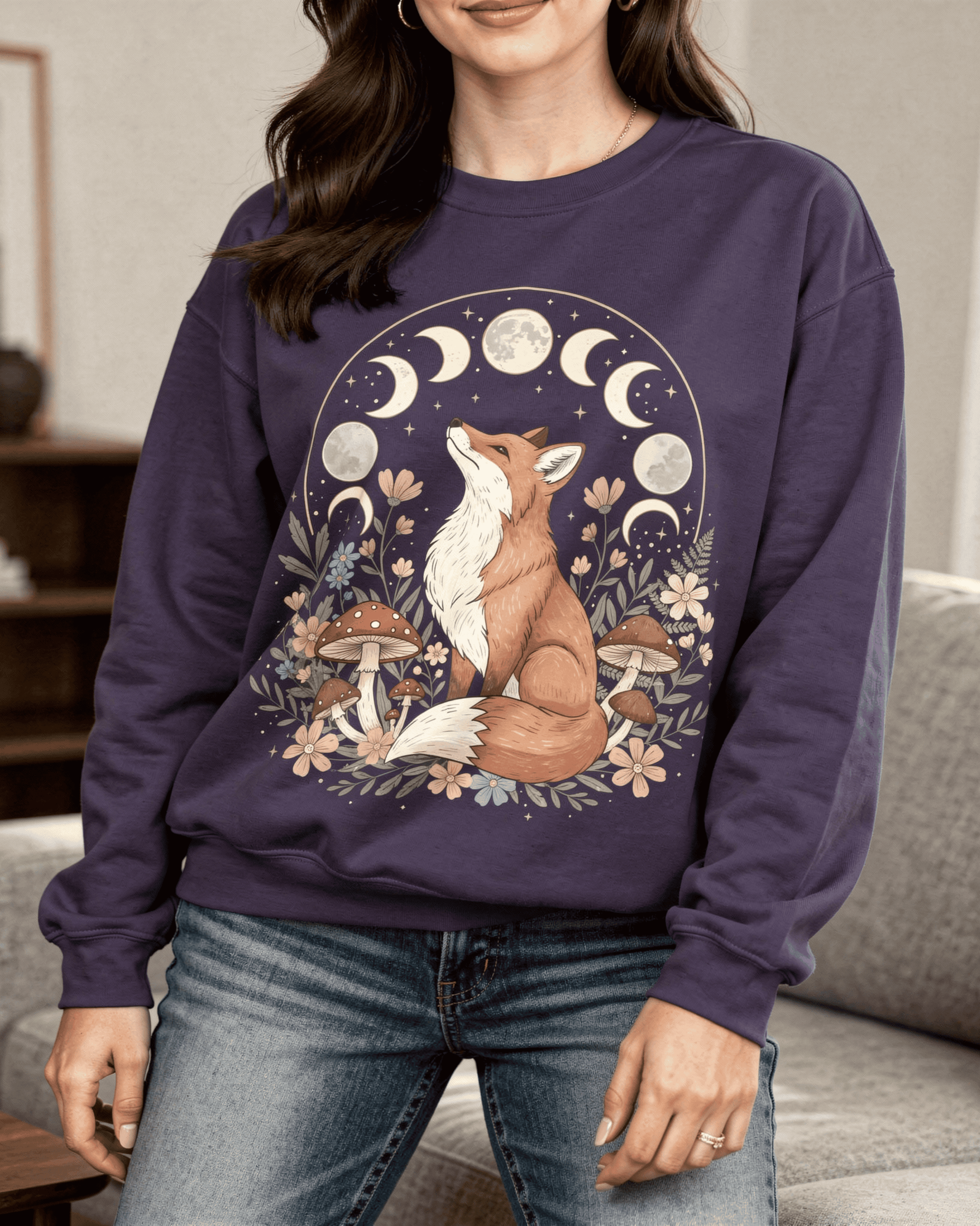 A woman models a dark purple sweatshirt featuring a beautiful cottagecore design. The graphic shows an illustrated red fox sitting among mushrooms and wildflowers, gazing up at the full cycle of the moon phases.