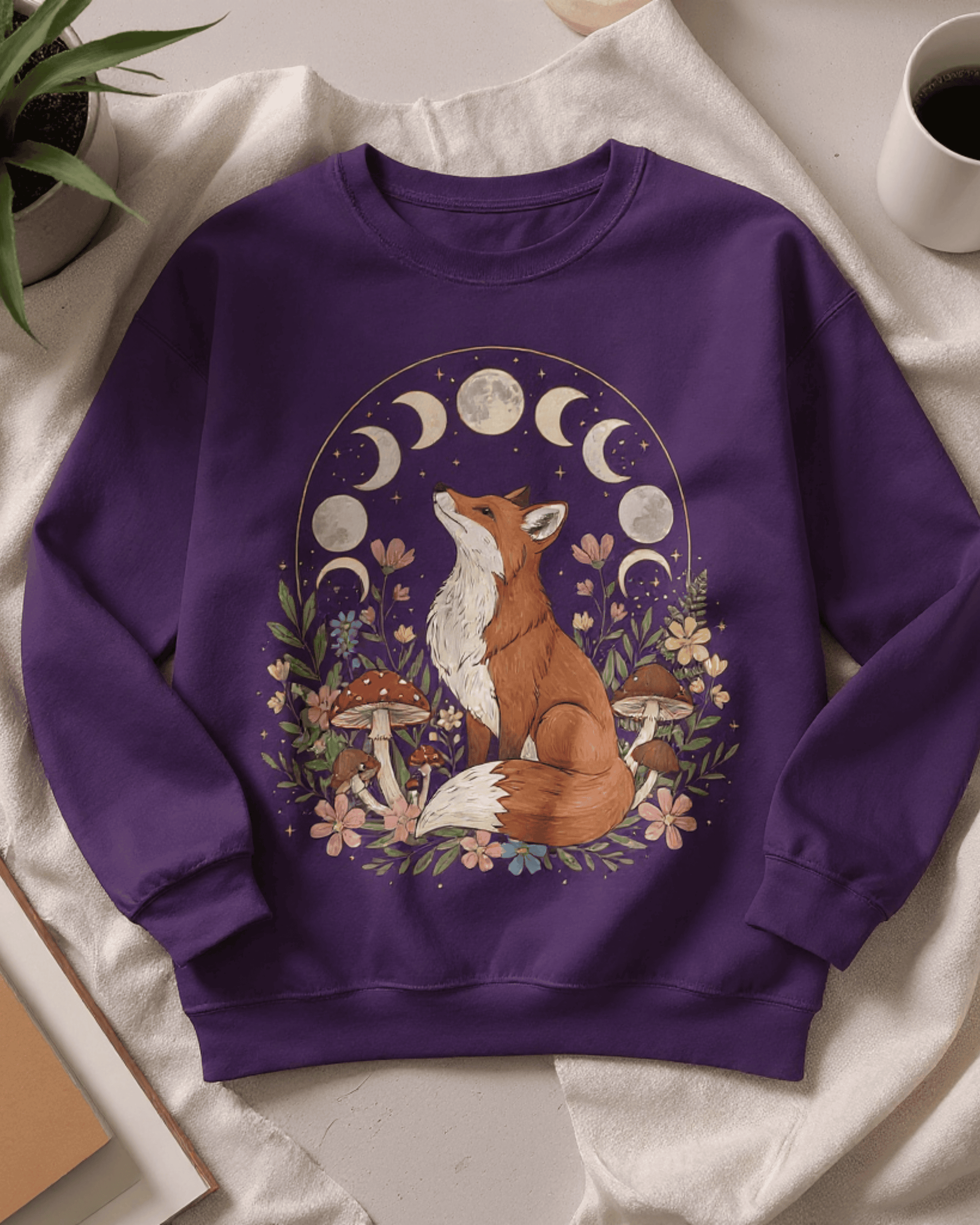 Close-up flat lay of the cozy cottagecore fox sweatshirt. The detailed graphic shows a red fox surrounded by botanicals, looking up at the different moon phases, all printed on a rich purple crewneck.