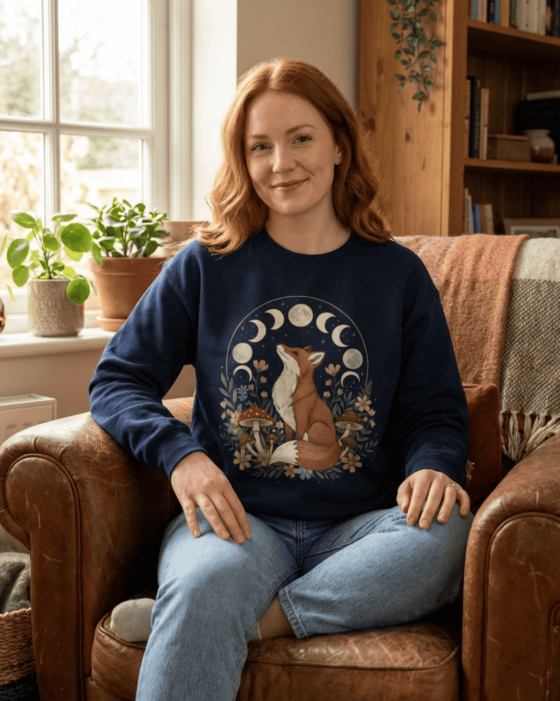 Woman relaxing in a cozy cottage living room wearing a navy cottagecore fox sweatshirt decorated with moon phases, wildflowers and toadstool mushrooms.
