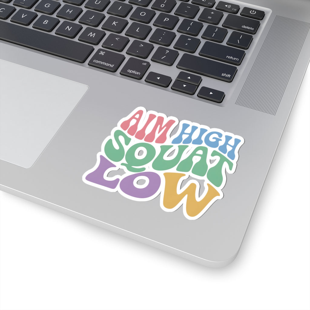 A colorful kiss-cut sticker with the text 'AIM HIGH SQUAT LOW' in pastel rainbow letters, shown applied to the silver palm rest of a laptop keyboard.