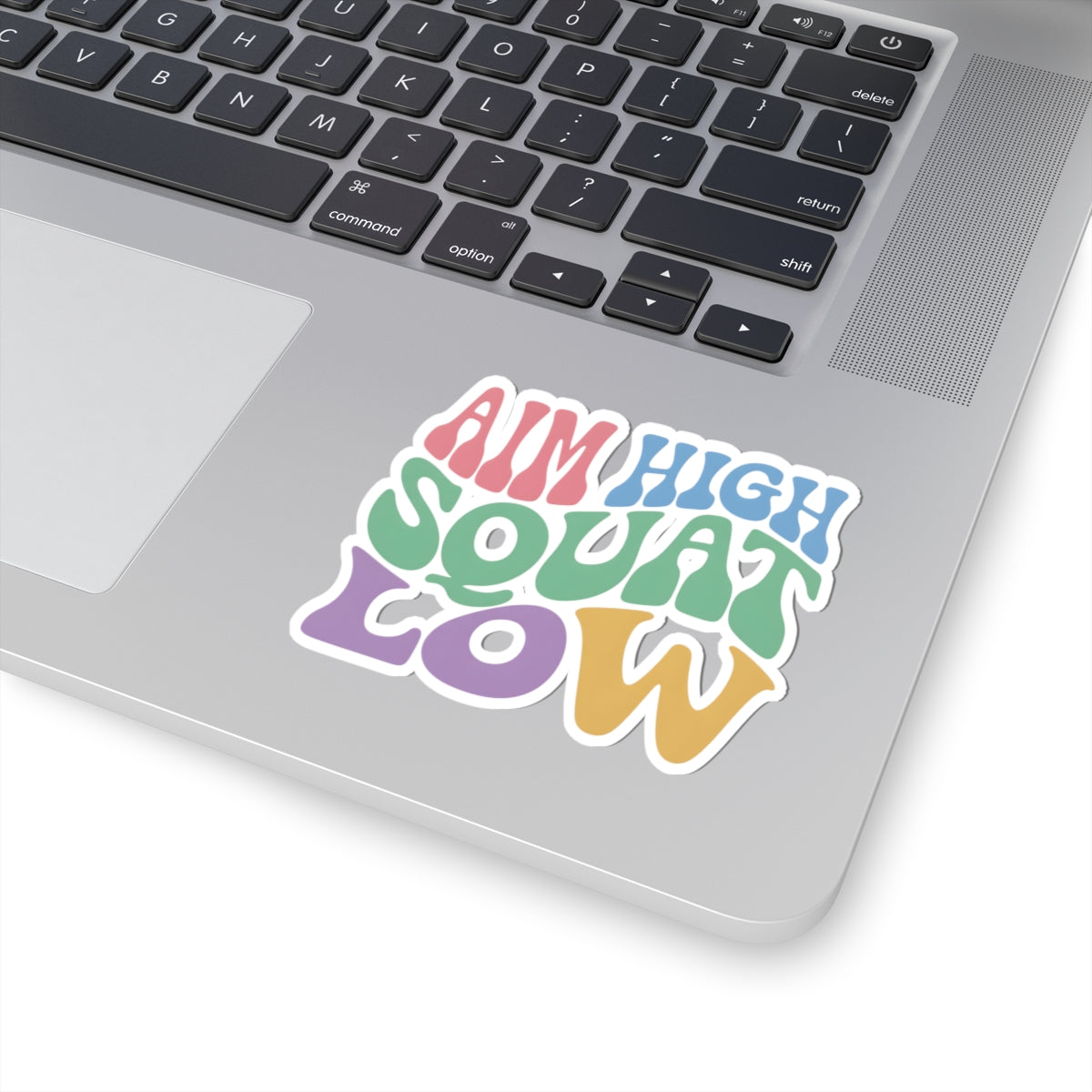 A colorful kiss-cut sticker with the text 'AIM HIGH SQUAT LOW' in pastel rainbow letters, shown applied to the silver palm rest of a laptop keyboard.