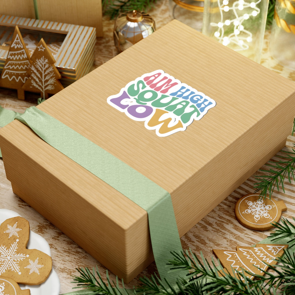 A kraft paper gift box tied with a pale green ribbon, featuring a colorful kiss-cut sticker that reads 'AIM HIGH SQUAT LOW' on top. The box is styled on a rustic wooden surface with holiday decorations including pine branches, gingerbread cookies, and another gift box in the background.