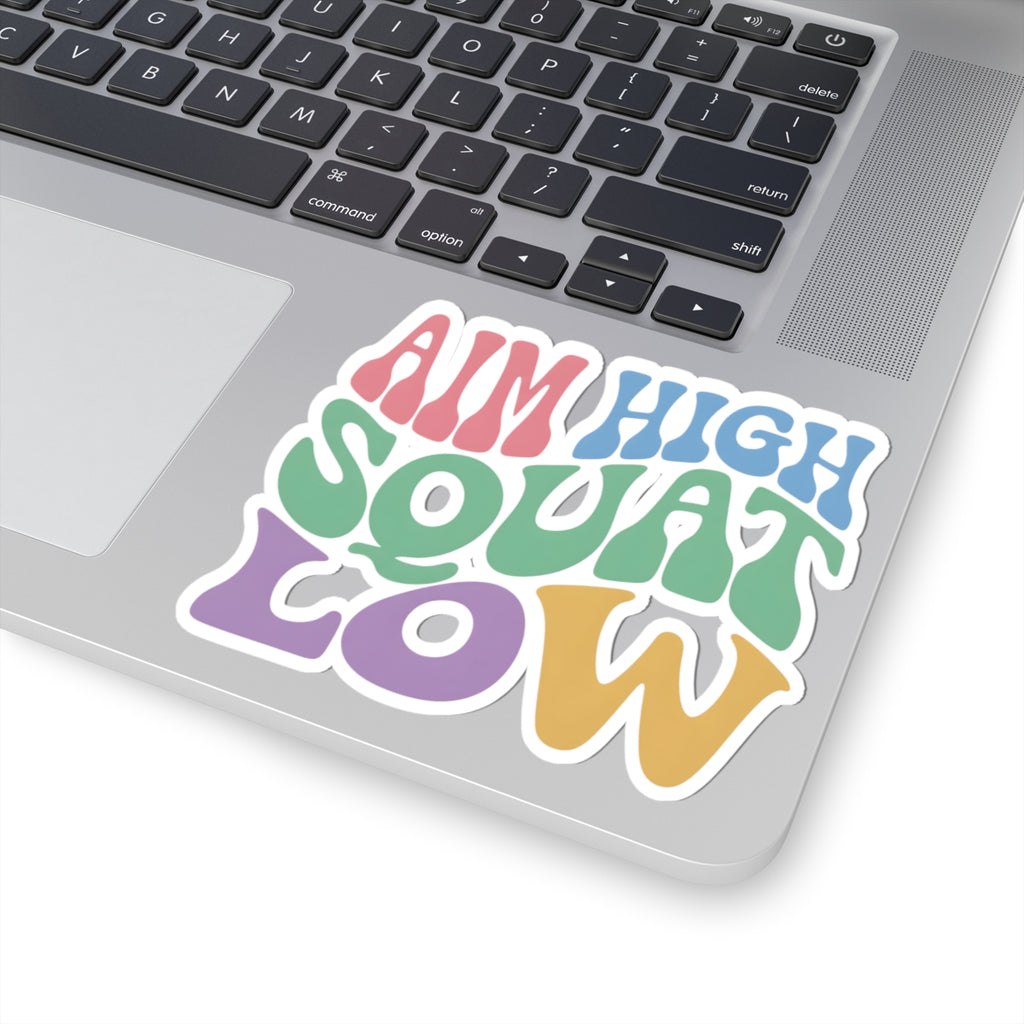 A colorful retro-style kiss-cut sticker reading 'AIM HIGH SQUAT LOW' in pink, blue, green, purple, and yellow lettering, shown applied to the silver surface of a laptop near the keyboard.