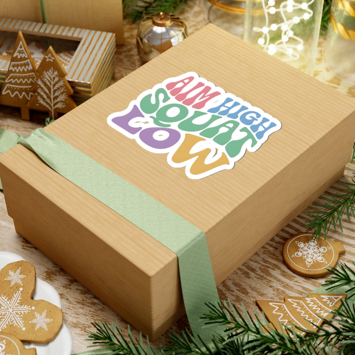 A square kraft paper gift box tied with a pale green ribbon, featuring a colorful kiss-cut sticker that reads “AIM HIGH SLOW” in rainbow letters. The box is styled on a rustic wooden surface with holiday decorations including gingerbread cookies, evergreen branches, and a small ornament.