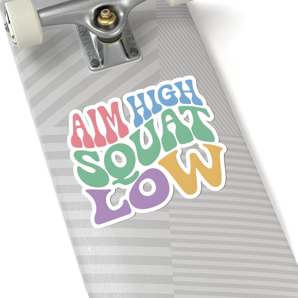 A close-up of a skateboard deck with a colorful retro-style sticker that reads 'AIM HIGH SQUAT LOW' in pastel pink, blue, green, yellow, and purple lettering. The skateboard has a gray and white striped pattern, and part of the silver truck and white wheels are visible.