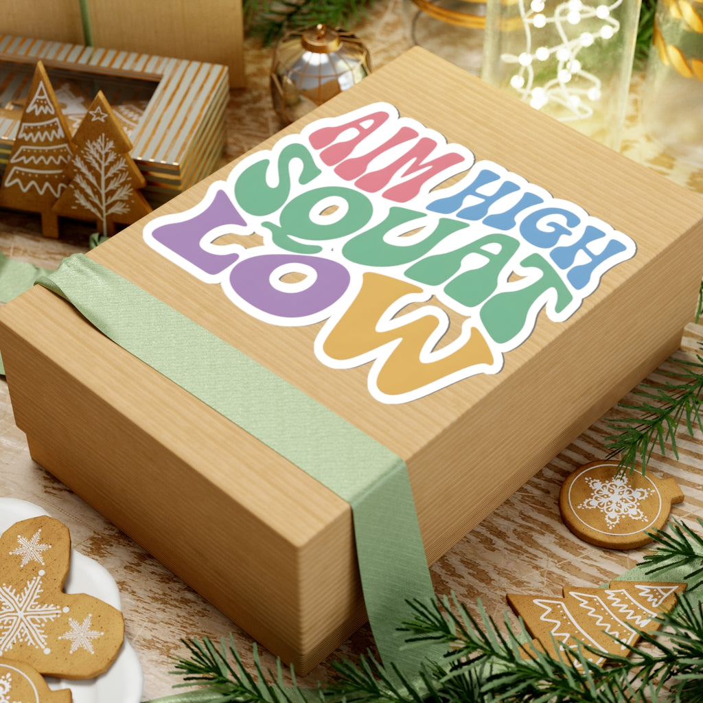 A wooden gift box wrapped with a sage green ribbon, featuring a colorful kiss-cut sticker that reads 'AIM HIGH SQUAT LOW' in pastel rainbow letters. The box is styled on a rustic wooden table with Christmas cookies, pine branches, and holiday decorations.