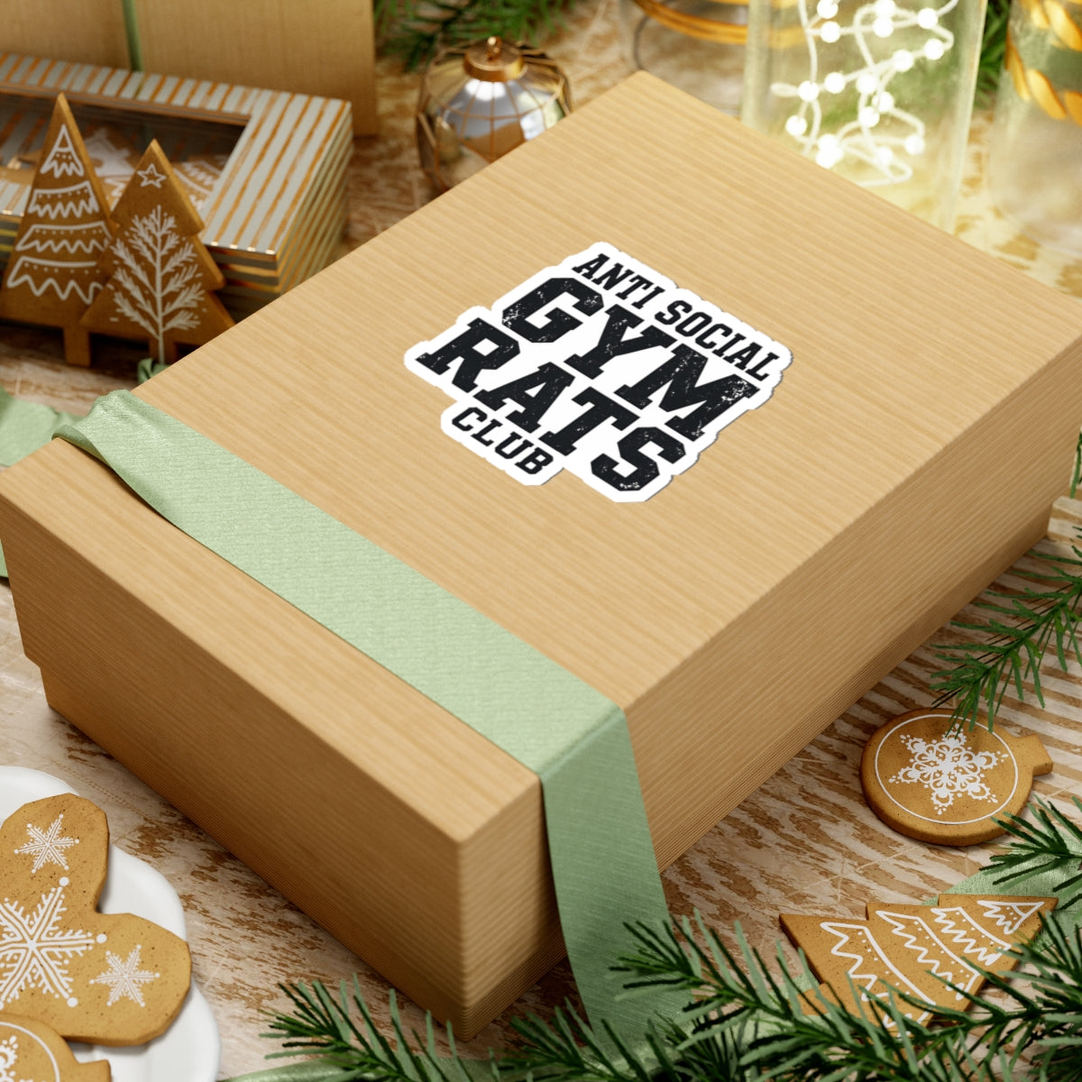 Wooden gift box with a green ribbon and 'Anti Social Gym Rats Club' sticker on a festive background with cookies and lights.