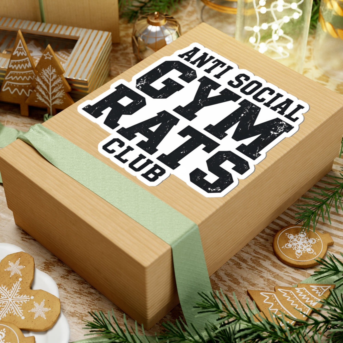 A rectangular kraft paper gift box wrapped with a pale green ribbon, featuring a large kiss-cut sticker that reads “ANTI SOCIAL GYM RATS CLUB” in bold black letters with a white outline. The box is styled on a wooden surface with holiday decorations, including gingerbread cookies, pine branches, and a small decorative box in the background.