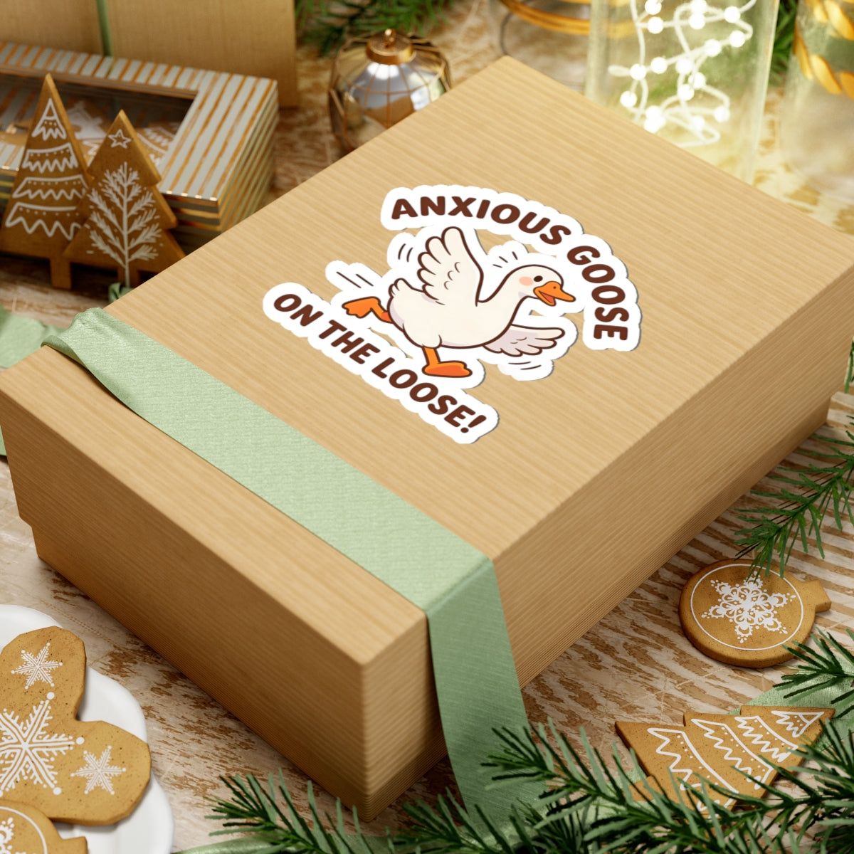 A kraft paper gift box tied with a pale green ribbon, featuring a kiss-cut sticker that reads 'ANXIOUS GOOSE ON THE LOOSE!' with a cartoon goose. The box is styled on a tabletop with holiday decorations, gingerbread cookies, and pine branches.