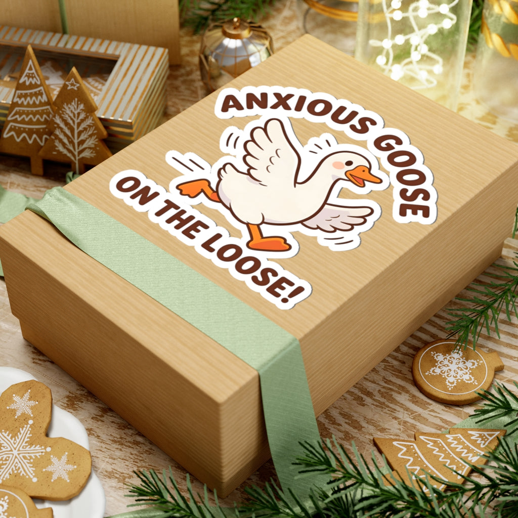 A light wood gift box wrapped with a pale green ribbon, featuring a kiss-cut sticker of a white goose with orange beak and feet and the text 'ANXIOUS GOOSE ON THE LOOSE!' on top. The box is styled with holiday cookies, evergreen branches, and other gift boxes in the background.