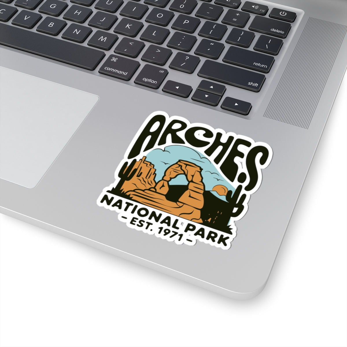 A kiss-cut sticker featuring a desert landscape with the words 'Arches National Park - Est. 1971' applied to the palm rest of a silver laptop.