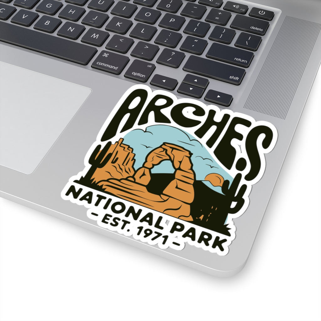 A kiss-cut vinyl sticker featuring a desert landscape illustration with the text 'ARCHES NATIONAL PARK - EST. 1971' applied to the corner of a silver laptop.