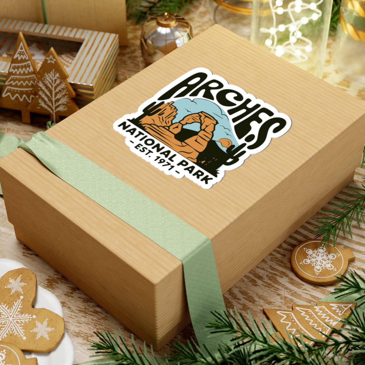 A square kraft gift box wrapped with a pale green ribbon, featuring a kiss-cut sticker that reads 'Arches National Park - Est. 1971' with a desert landscape illustration.