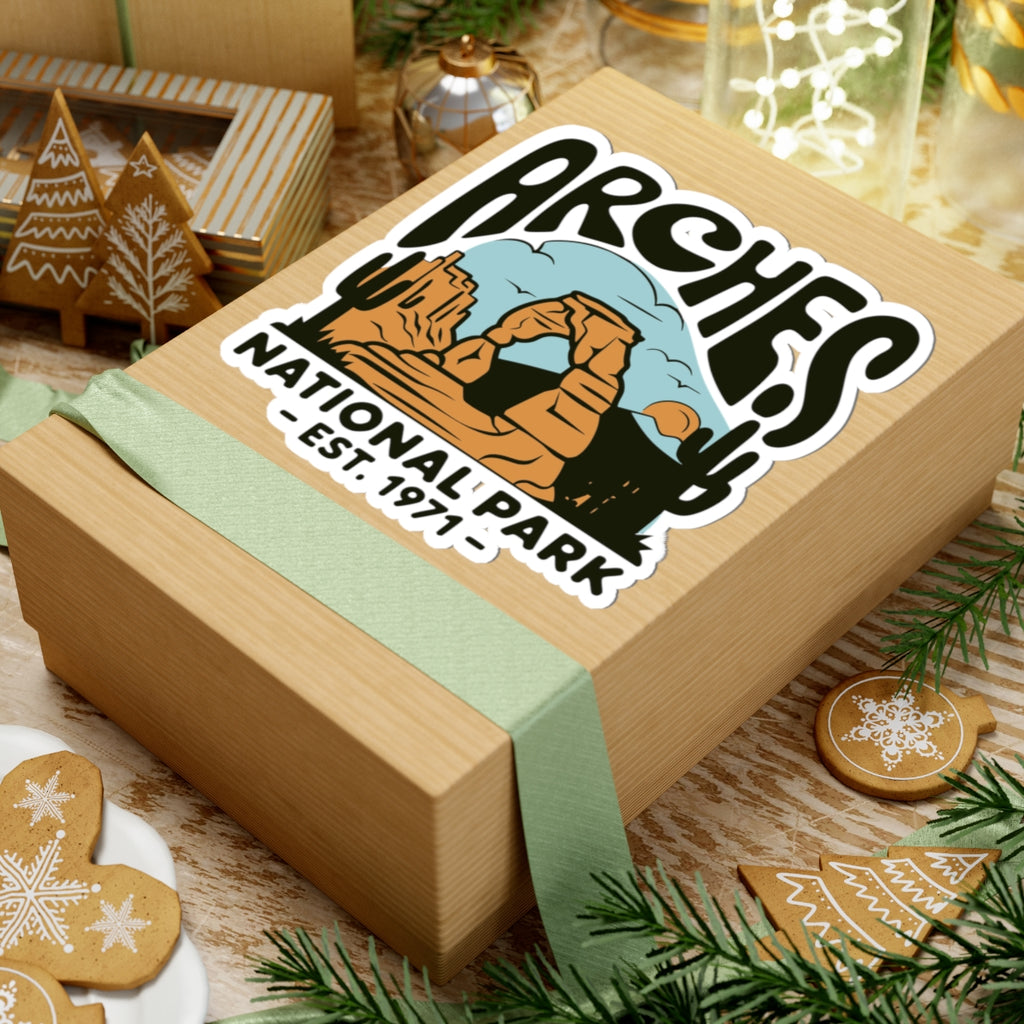 A wooden gift box wrapped with a pale green ribbon, featuring a large Arches National Park sticker on top, styled with holiday cookies, pine branches, and warm background decor.
