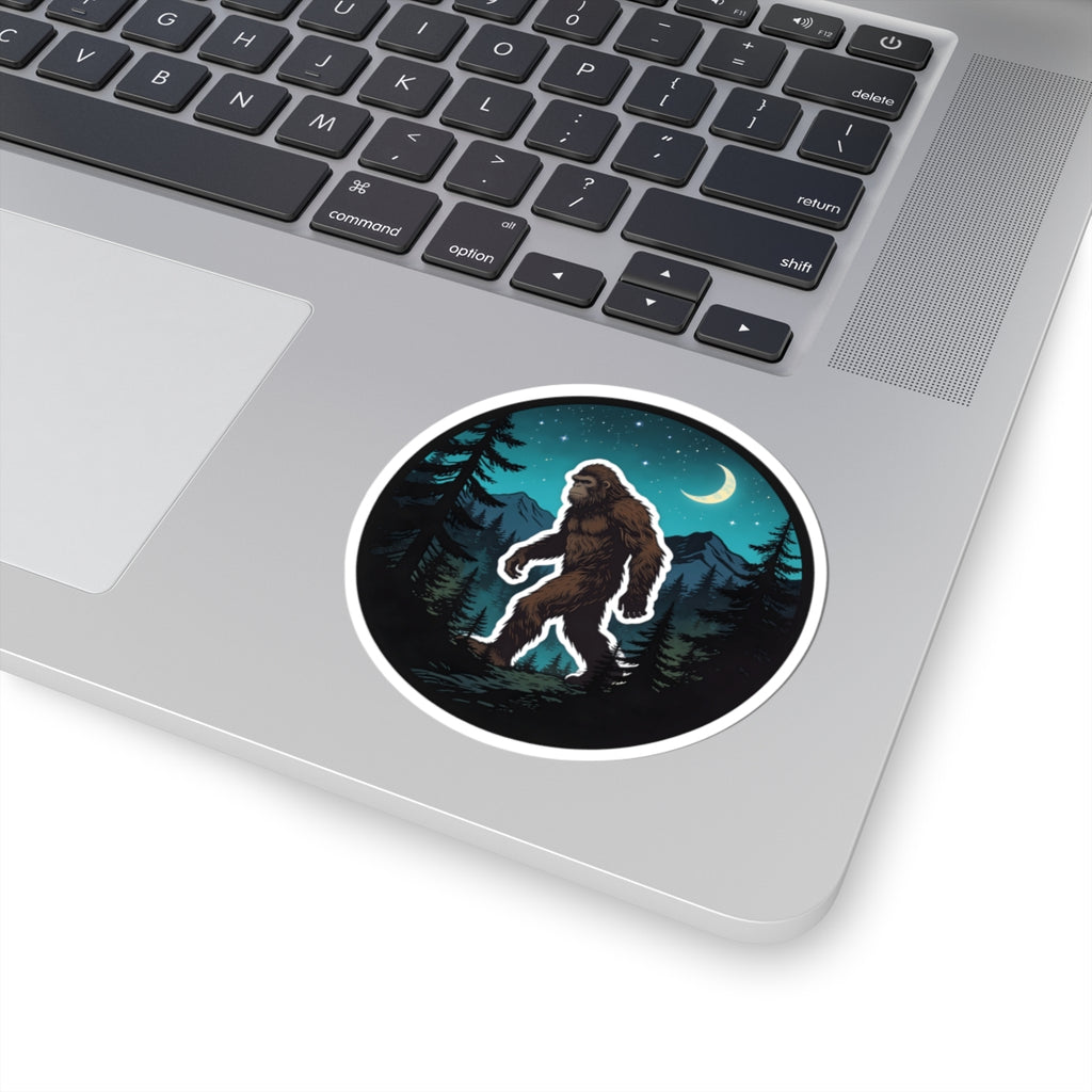 A round sticker with a cartoon Bigfoot in a night forest scene, shown applied to the corner of a silver laptop on a white background.