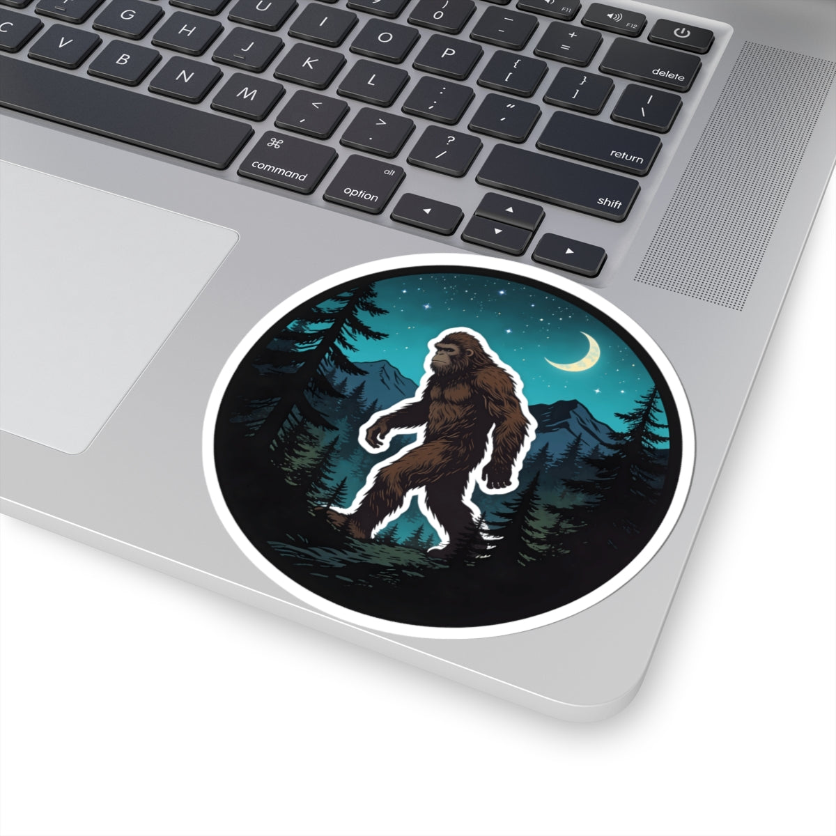A round sticker with a white border is applied to the palm rest of a silver laptop. The sticker features an illustration of a Bigfoot figure walking through a pine forest at night under a crescent moon.