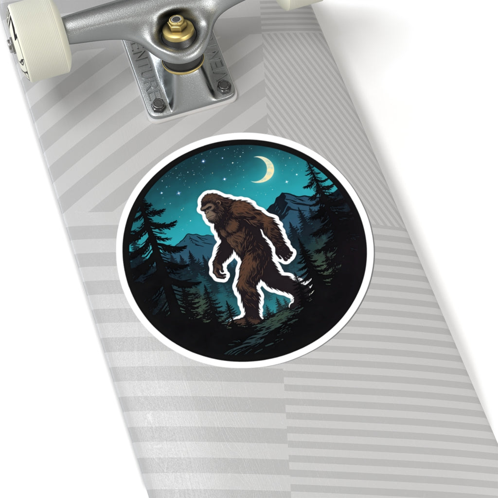 A close-up of a skateboard deck with a circular sticker featuring an illustrated Bigfoot in a dark forest at night under a crescent moon.