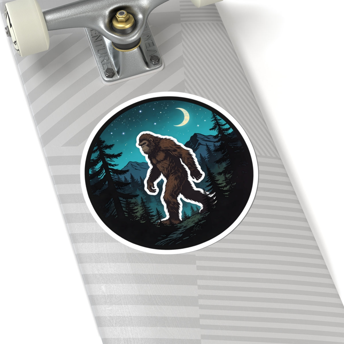 A close-up of a skateboard deck with a circular sticker featuring an illustrated Bigfoot in a dark forest at night under a crescent moon.
