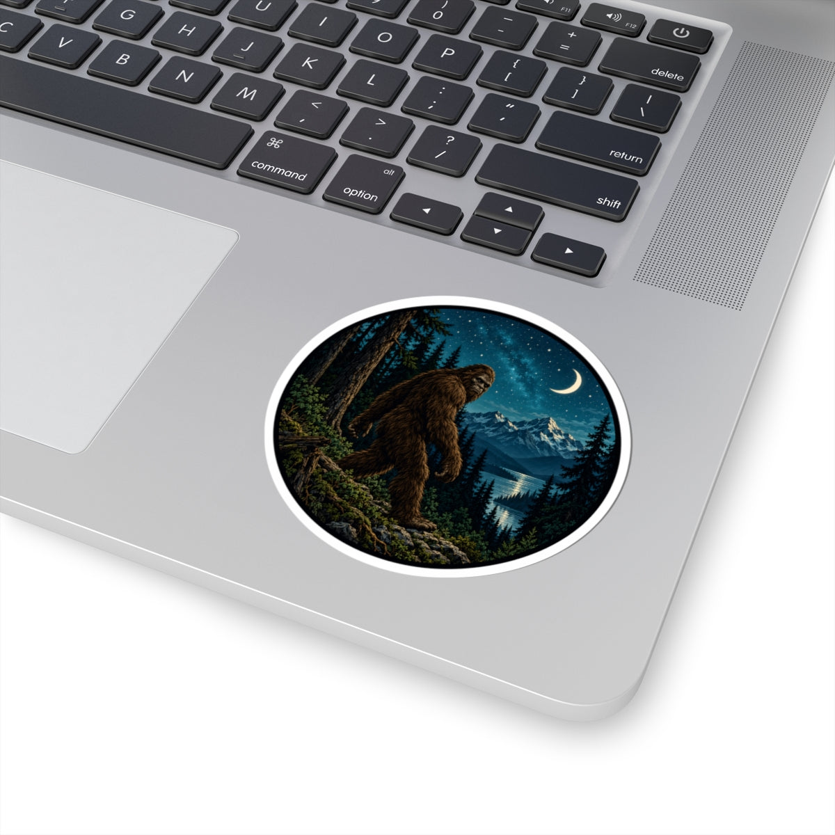 A round vinyl sticker with a white border is applied to the palm rest of a silver laptop. The sticker features an illustration of a Bigfoot figure standing in a dark pine forest at night under a crescent moon and starry sky.