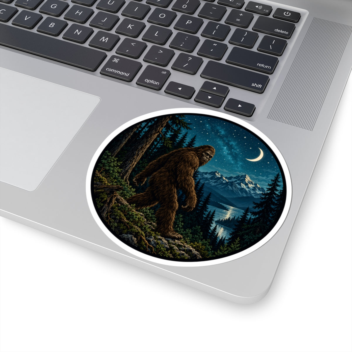 A round vinyl sticker with a white border is shown applied to the lower-left corner of a silver laptop. The sticker features an illustration of a Bigfoot figure standing in a dark pine forest at night under a starry sky and crescent moon, with snow-capped mountains and a reflective lake in the distance.