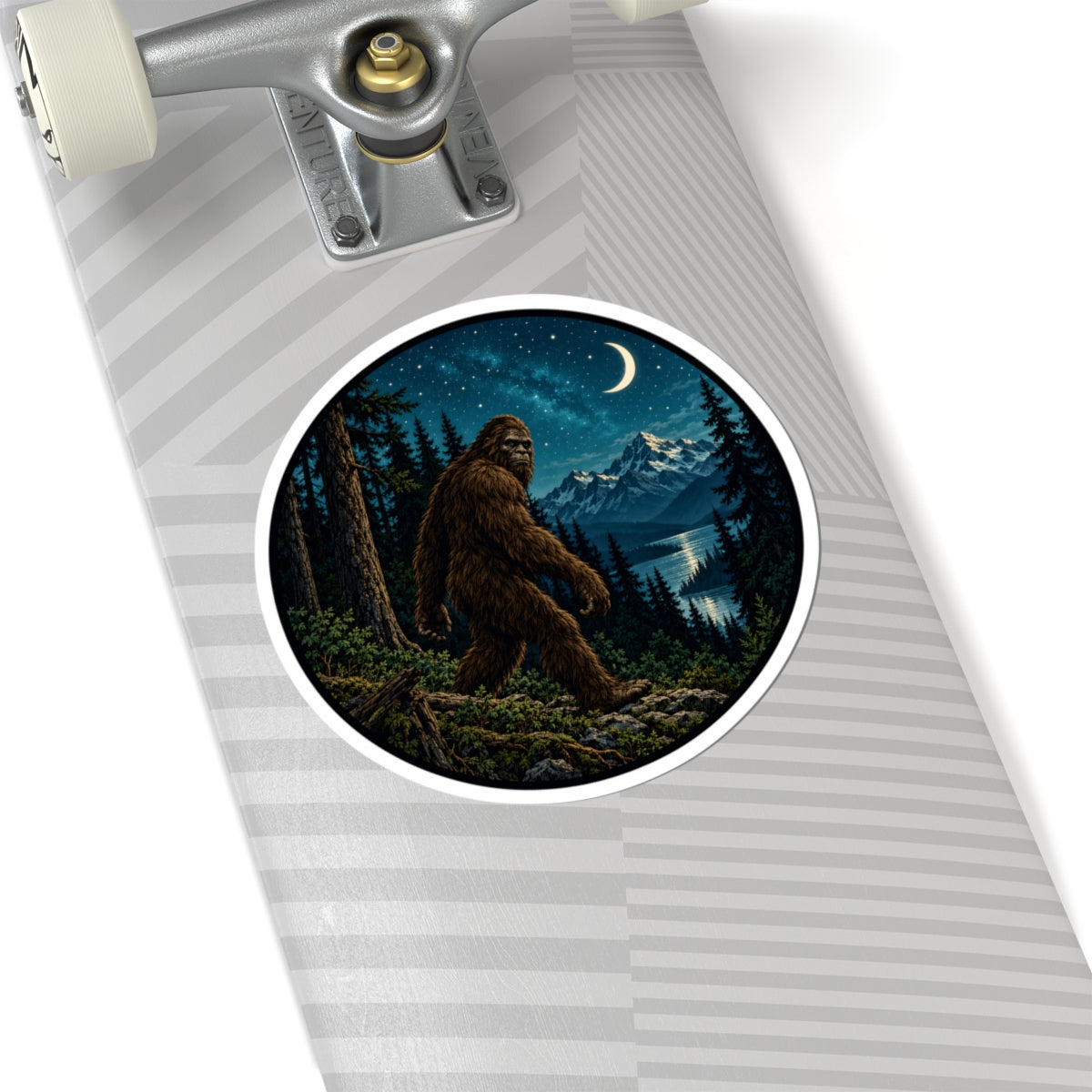 A close-up of a skateboard deck featuring a circular sticker of Bigfoot in a forest at night under a crescent moon, with a silver trucks and white wheels visible of the skateboard. 