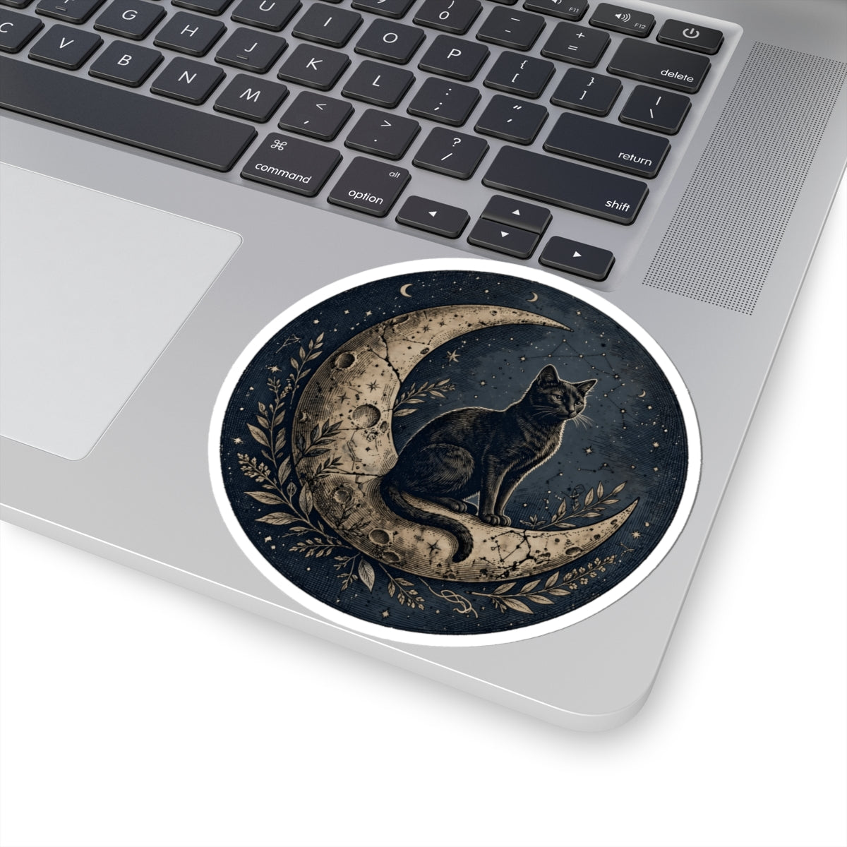 A round sticker with a gothic illustration of a black cat sitting on a crescent moon, placed on the palm rest of a silver laptop.