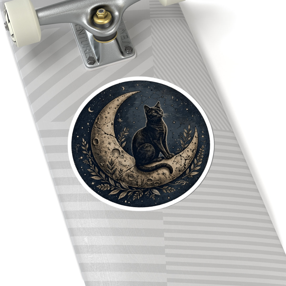 A close-up of a skateboard deck featuring a circular sticker of a black cat sitting on a crescent moon under a starry night sky, with a gray striped deck background and part of the silver truck visible.