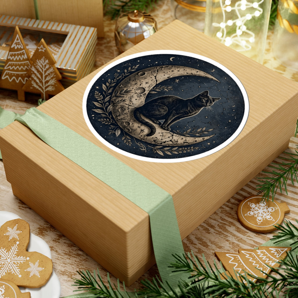 A square kraft gift box wrapped with a pale green ribbon, topped with a round sticker featuring a black cat sitting on a crescent moon with a tree branch motif. The box is styled on a tabletop with holiday cookies, evergreen branches, and other gift items in the background.
