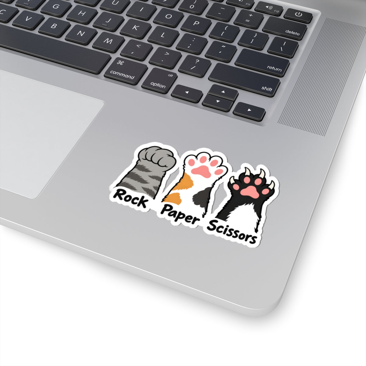 A close-up of a silver laptop showing a kiss-cut sticker on the palm rest. The sticker features three cat paw prints labeled 'Rock', 'Paper', and 'Scissors' in a classic scissors game design.