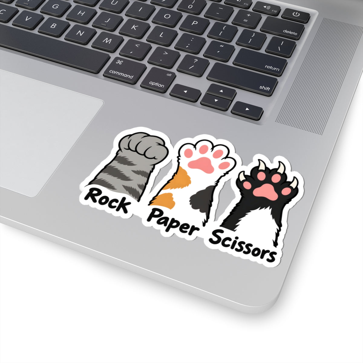 A close-up of a silver laptop showing a kiss-cut sticker on the palm rest. The sticker features three stylized cat paw prints labeled 'Rock', 'Paper', and 'Scissors' in gray, orange/black, and black/white, with black text below.
