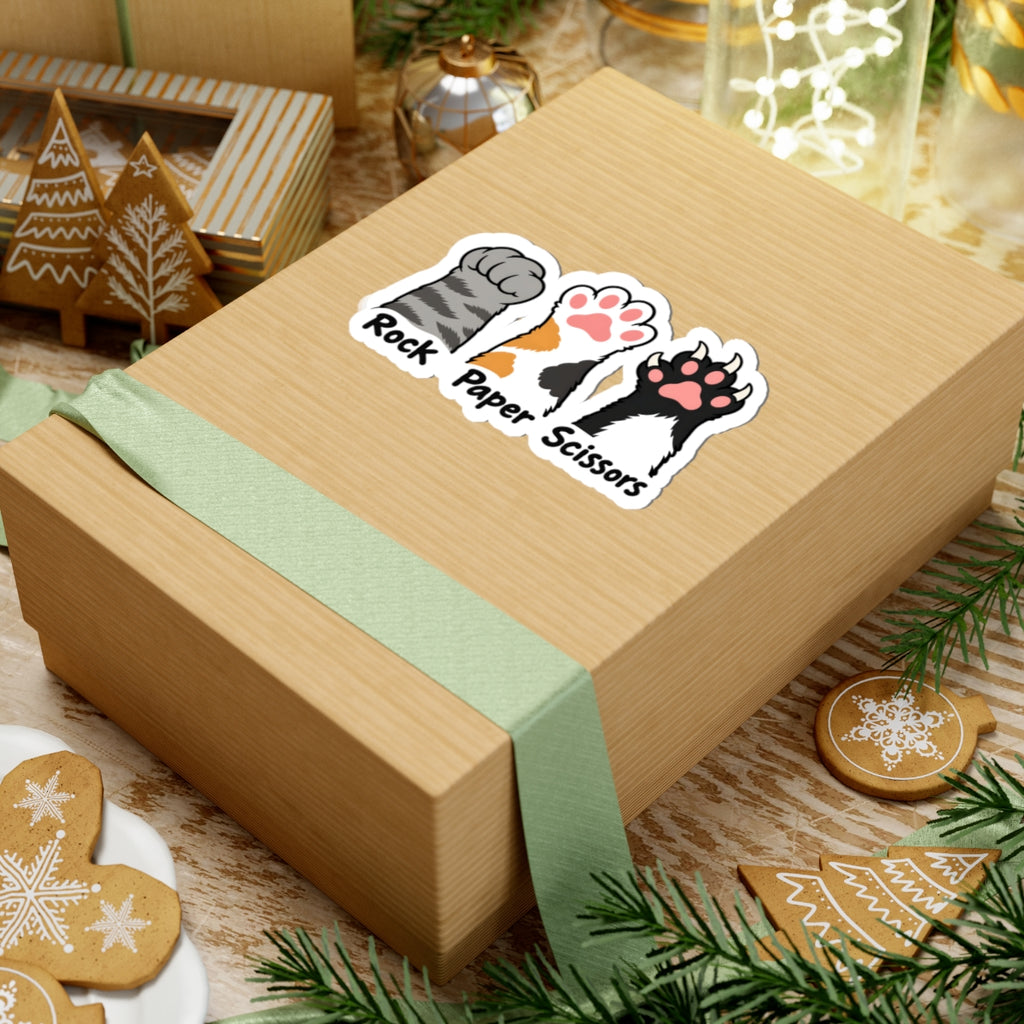 A kraft paper gift box tied with a pale green ribbon, decorated with a kiss-cut sticker that reads 'Rock Paper Scissors' with illustrated cat paw prints. The box is styled on a wooden surface with holiday decorations including gingerbread cookies, evergreen branches, and glass jars.
