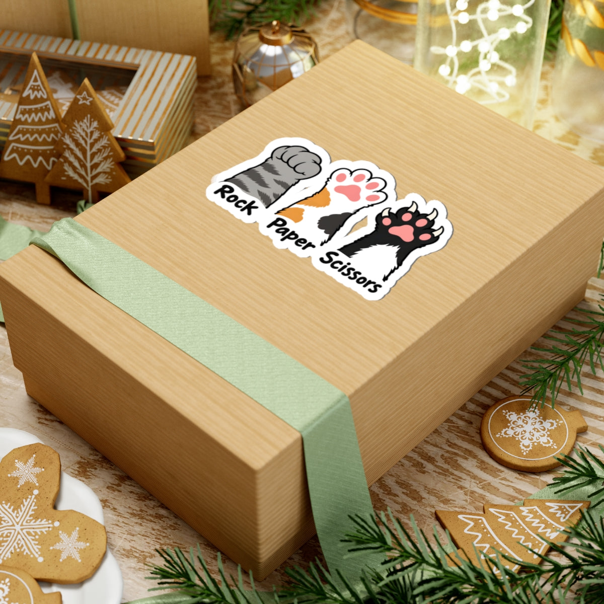 A kraft paper gift box tied with a pale green ribbon, decorated with a kiss-cut sticker that reads 'Rock Paper Scissors' with illustrated cat paw prints. The box is styled on a wooden surface with holiday decorations including gingerbread cookies, evergreen branches, and glass jars.