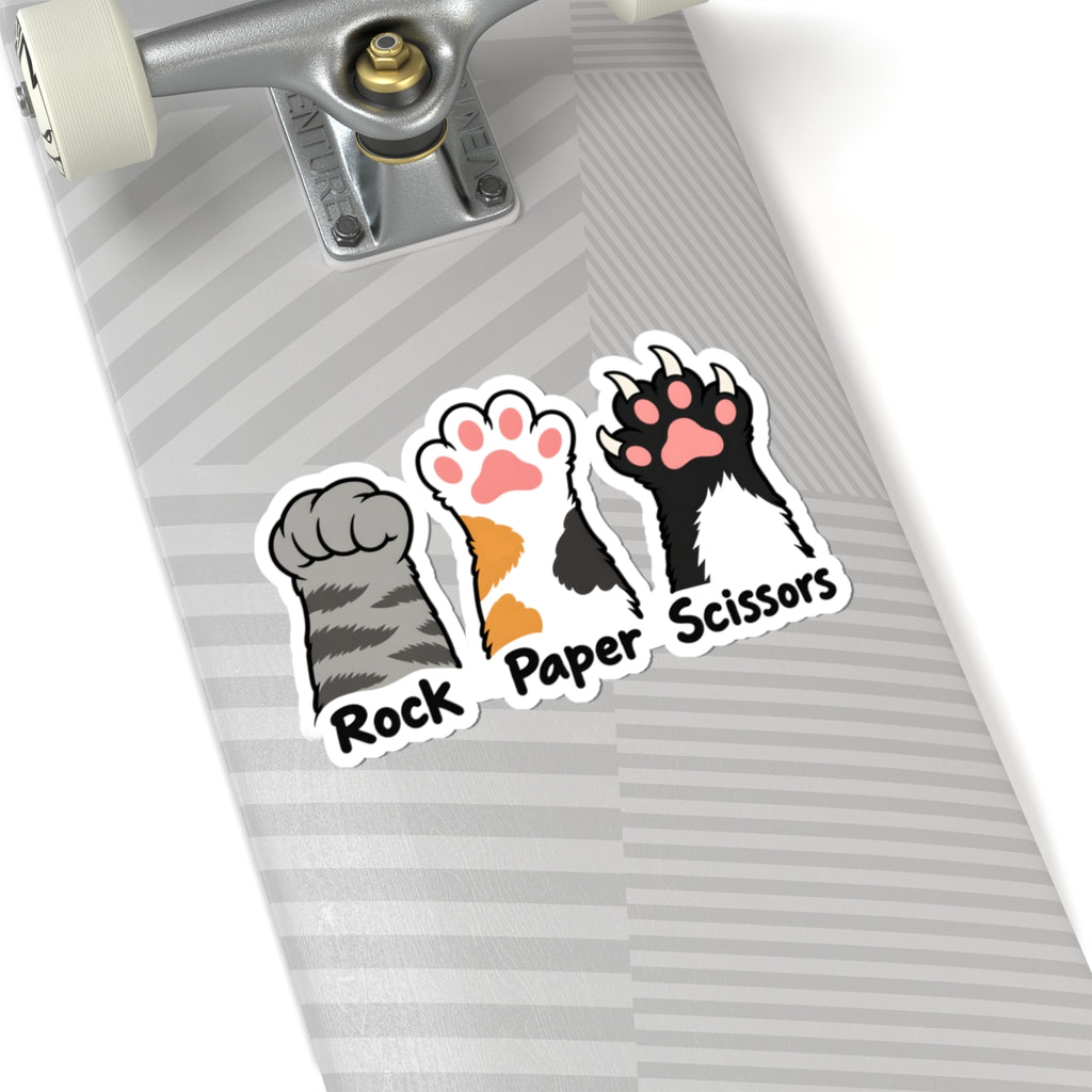 A close-up of a skateboard deck featuring three kiss-cut stickers of cat paw prints labeled 'Rock', 'Paper', and 'Scissors' in a rock-paper-scissors style. The skateboard has a gray and white striped pattern and is shown with part of the silver truck and white wheels visible against a clean white background.
