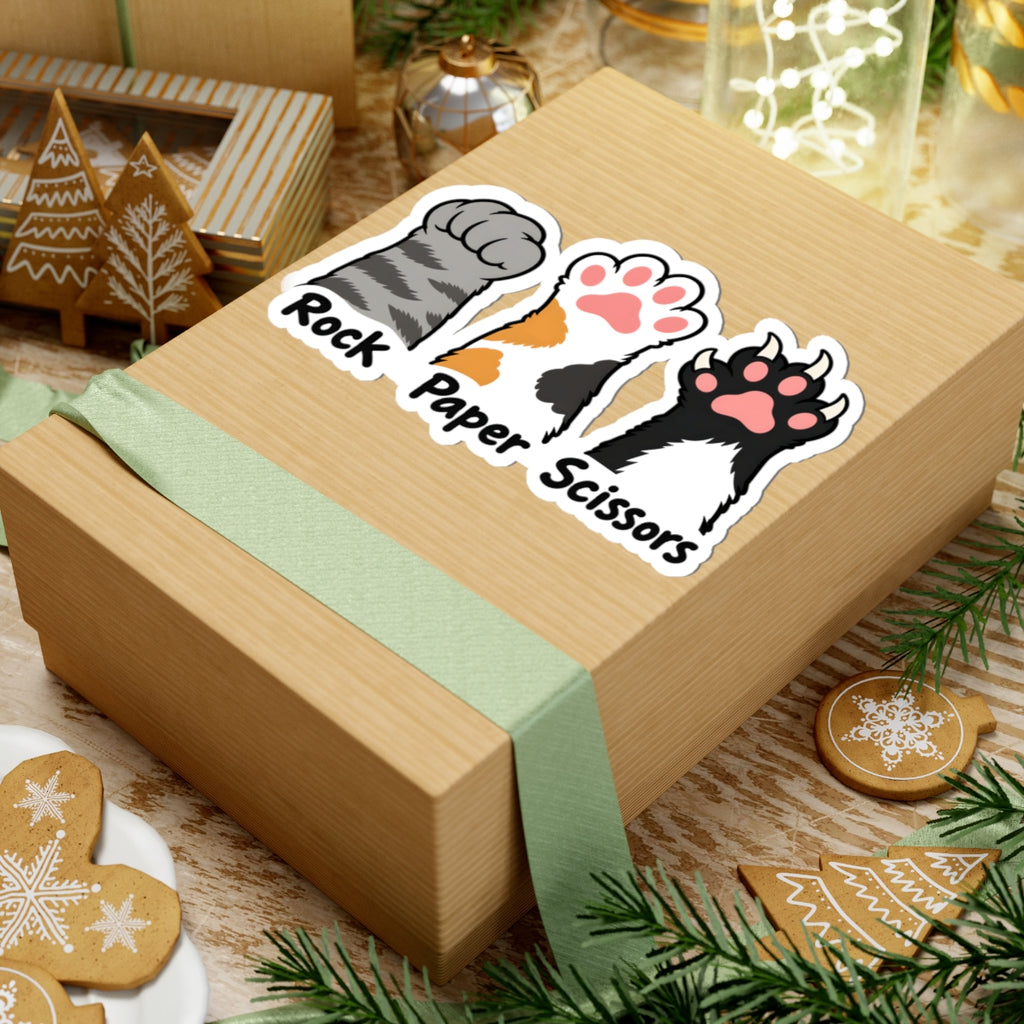 A square kraft paper gift box wrapped with a pale green ribbon, featuring a kiss-cut sticker of three cat paws labeled 'Rock Paper Scissors' on top. The box is styled on a wooden surface with holiday decorations including gingerbread cookies, pine branches, and a small tree ornament.