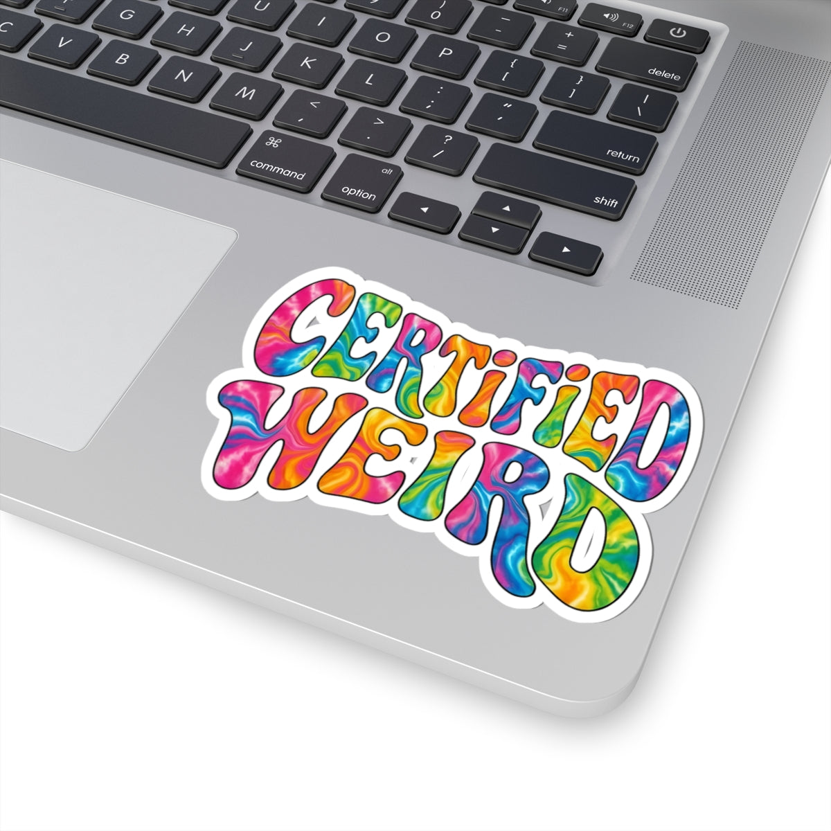 A kiss-cut vinyl sticker with the words 'CERTIFIED WEIRD' in a colorful tie-dye pattern, shown applied to the lower corner of a silver laptop keyboard.