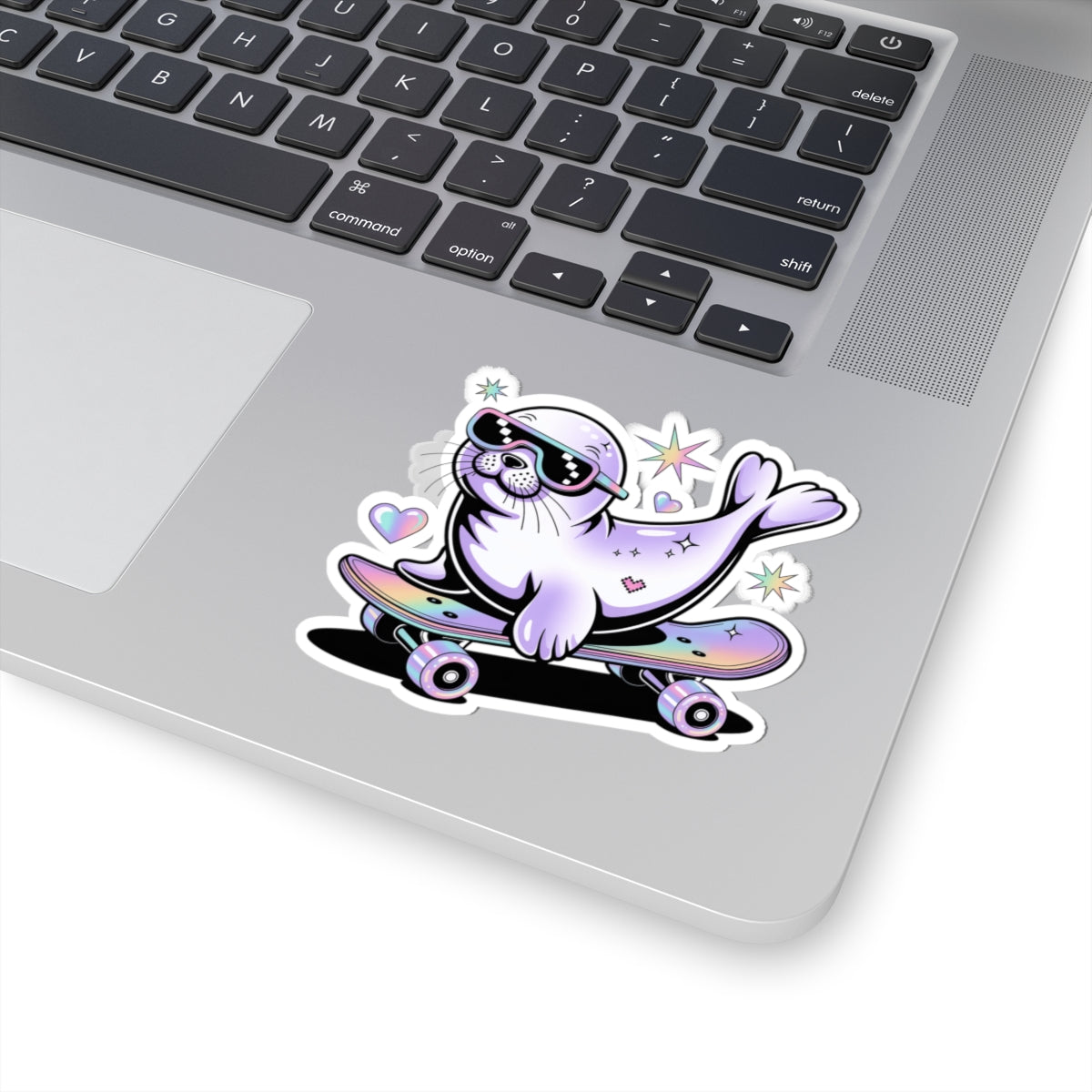 A kiss-cut vinyl sticker of a cute cartoon seal riding a skateboard, shown applied to the corner of a silver laptop. The sticker features a lavender seal wearing black sunglasses, with pastel pink, purple, and blue accents on the skateboard and small star and heart details.