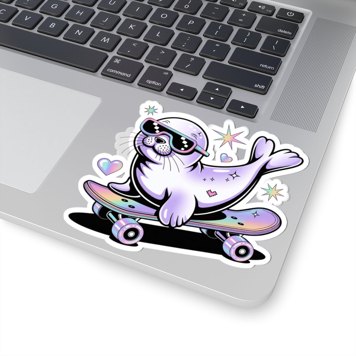 A kiss-cut vinyl sticker of a cartoon seal riding a skateboard, shown applied to the corner of a silver laptop. The sticker features a purple seal wearing black sunglasses, with pastel pink, blue, and purple accents on the skateboard and surrounding hearts and stars.