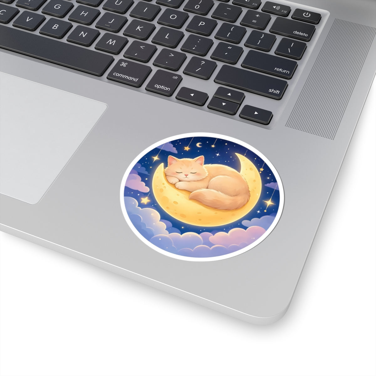 A round sticker of a sleeping orange cat on a crescent moon under a starry night sky, shown applied to the silver surface of a laptop next to the keyboard.