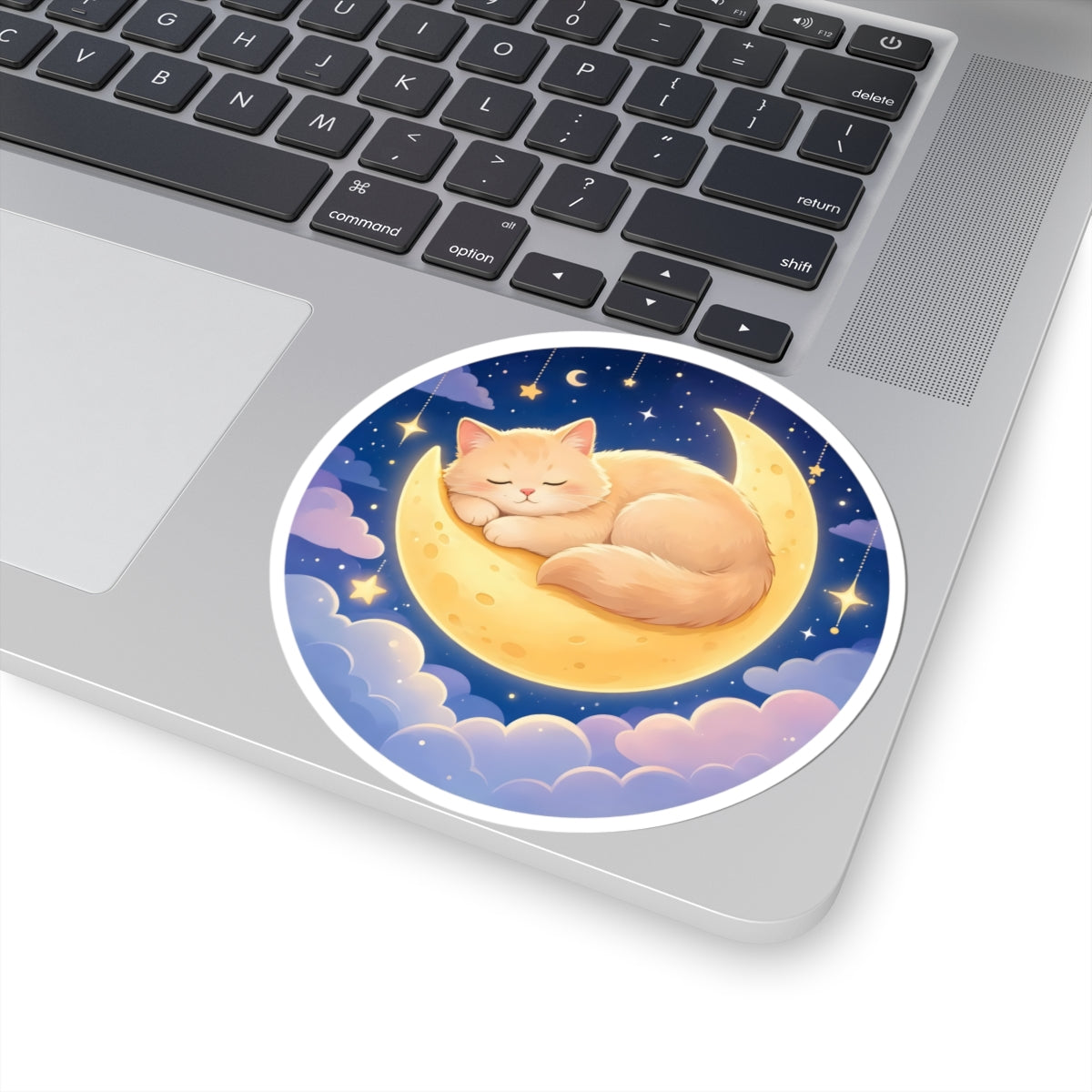 A round vinyl sticker of a sleeping orange tabby cat on a crescent moon, applied to the palm rest of a silver laptop.