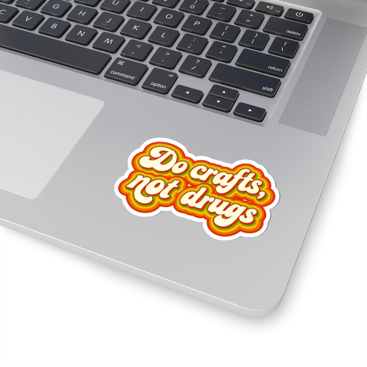A kiss-cut vinyl sticker with the text 'Do crafts, not drugs' in a retro bubble font, shown applied to the palm rest of a silver laptop. The sticker features a white background with a thick yellow and orange border.
