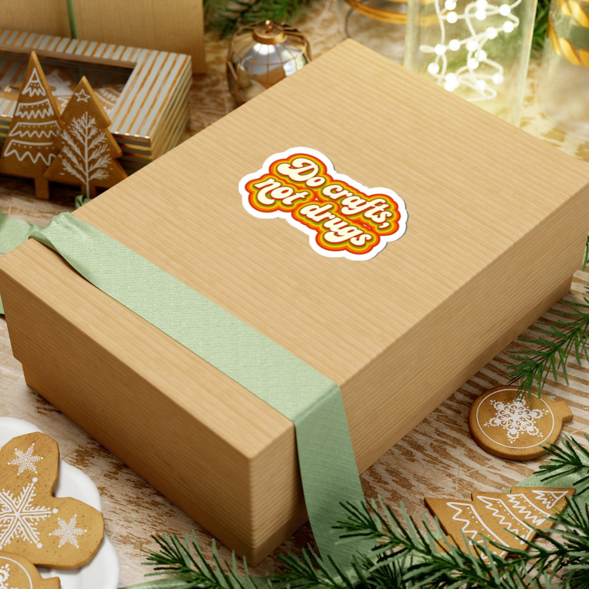 Wooden gift box with a green ribbon and a 'do crafts, not drugs' sticker on a festive background with cookies and lights.


