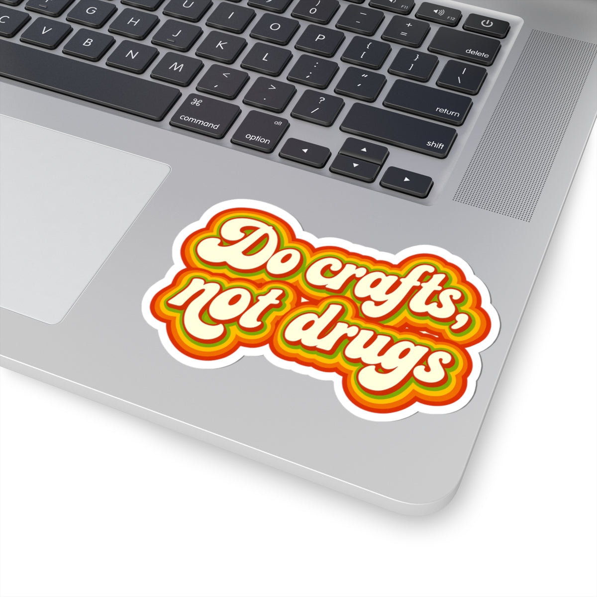 A retro-style kiss-cut sticker reading 'Do crafts, not drugs' in white script with yellow, orange, and red concentric outlines, shown applied to the lower area of a silver laptop keyboard.
