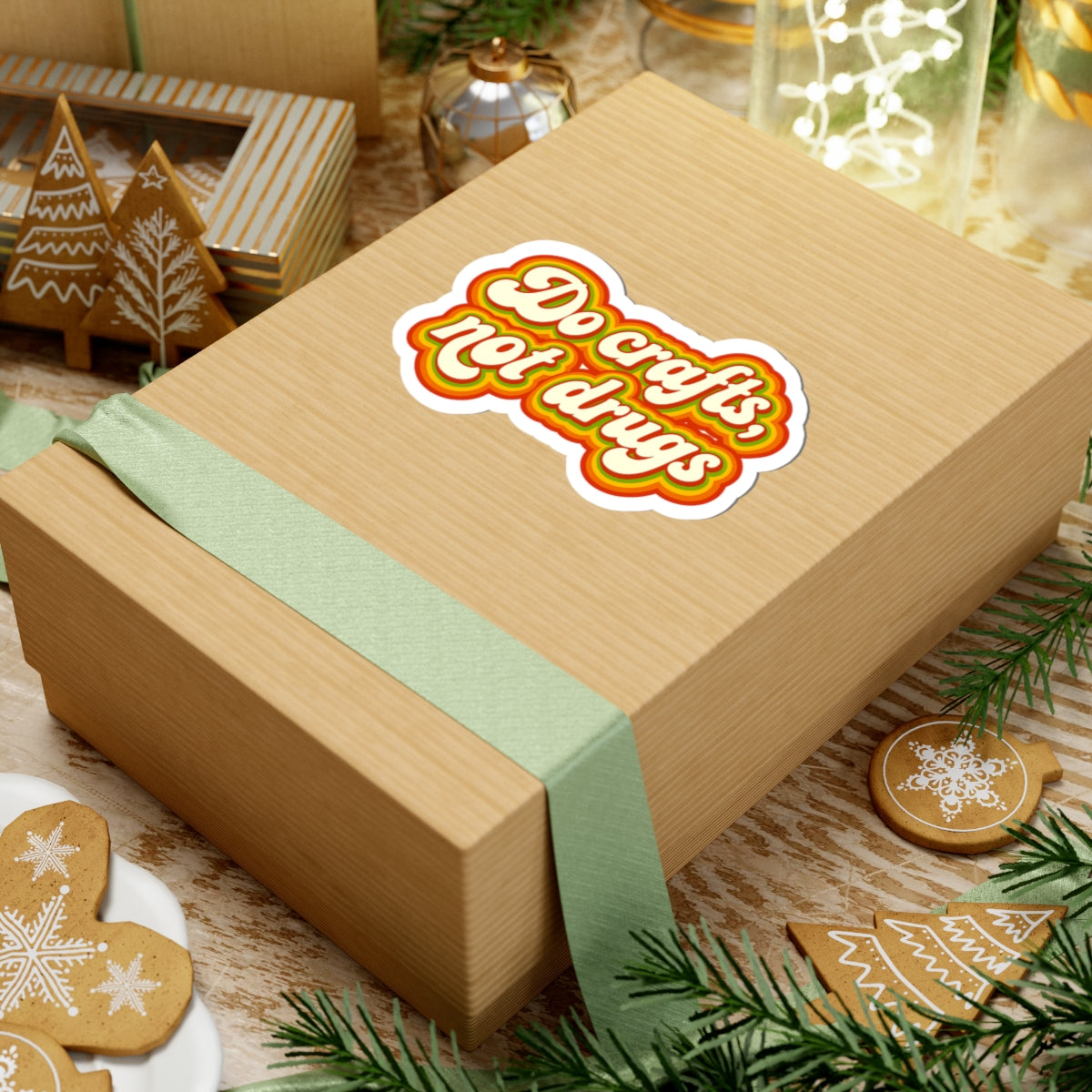 Wooden gift box with a green ribbon and a 'do crafts, not drugs' sticker on a festive background with cookies and lights.