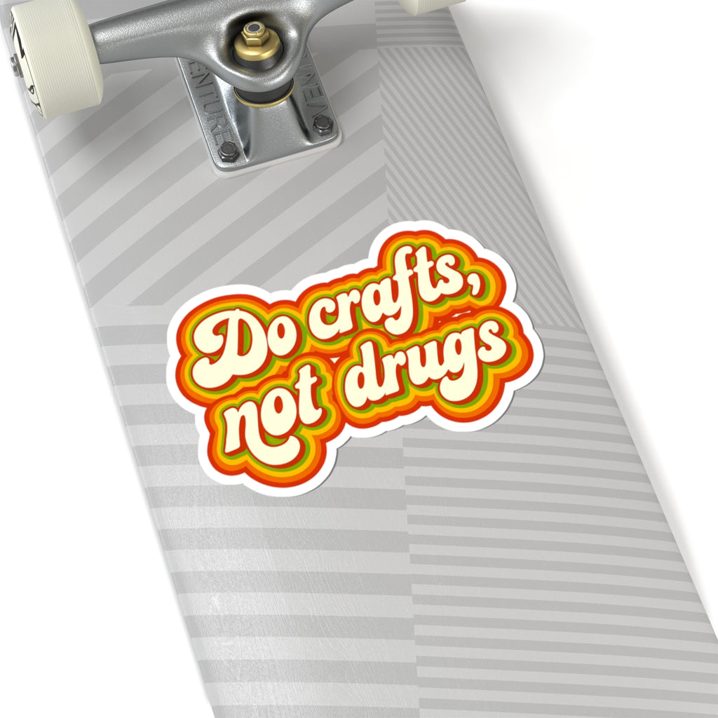A kiss-cut sticker with the text 'Do crafts, not drugs' in white retro lettering with yellow, orange, and red outlines, shown on a gray striped skateboard deck.