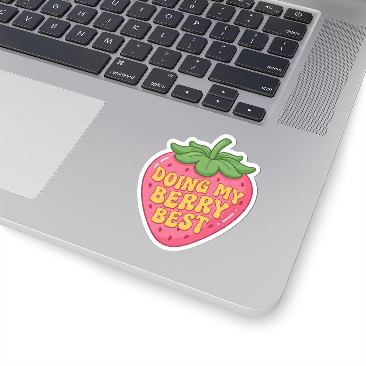 A strawberry-shaped sticker with the text 'DOING MY BERRY BEST' in yellow letters, shown applied to the palm rest of a silver laptop. The sticker has a pink body, green leafy top, and a white border.