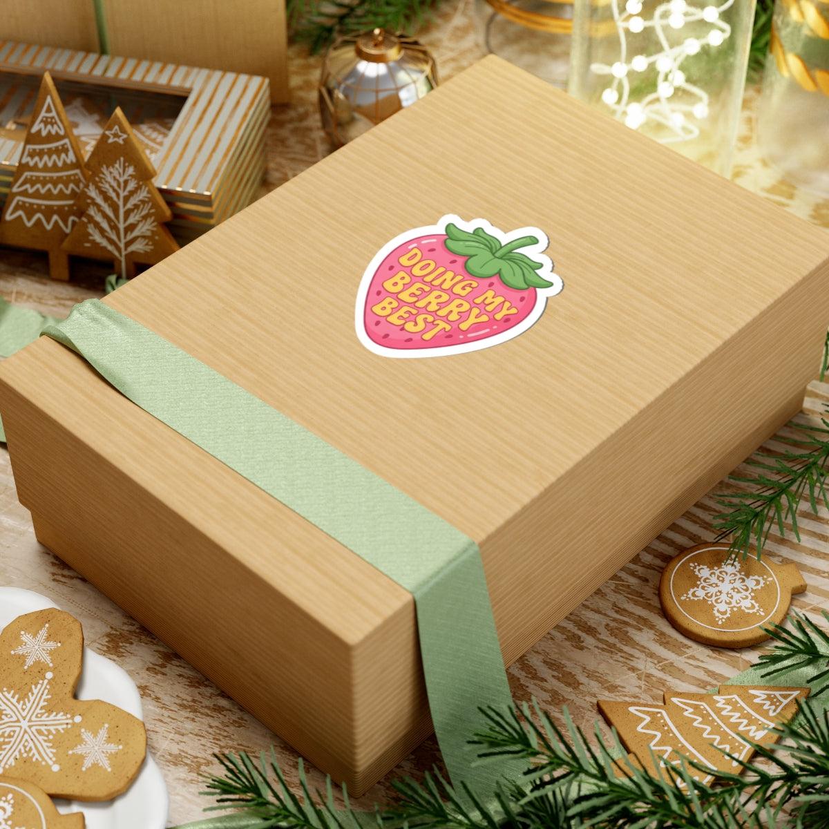 Gift box with a strawberry sticker with the text 'DOING MY BERRY BEST' on it, on a festive background with cookies and greenery.