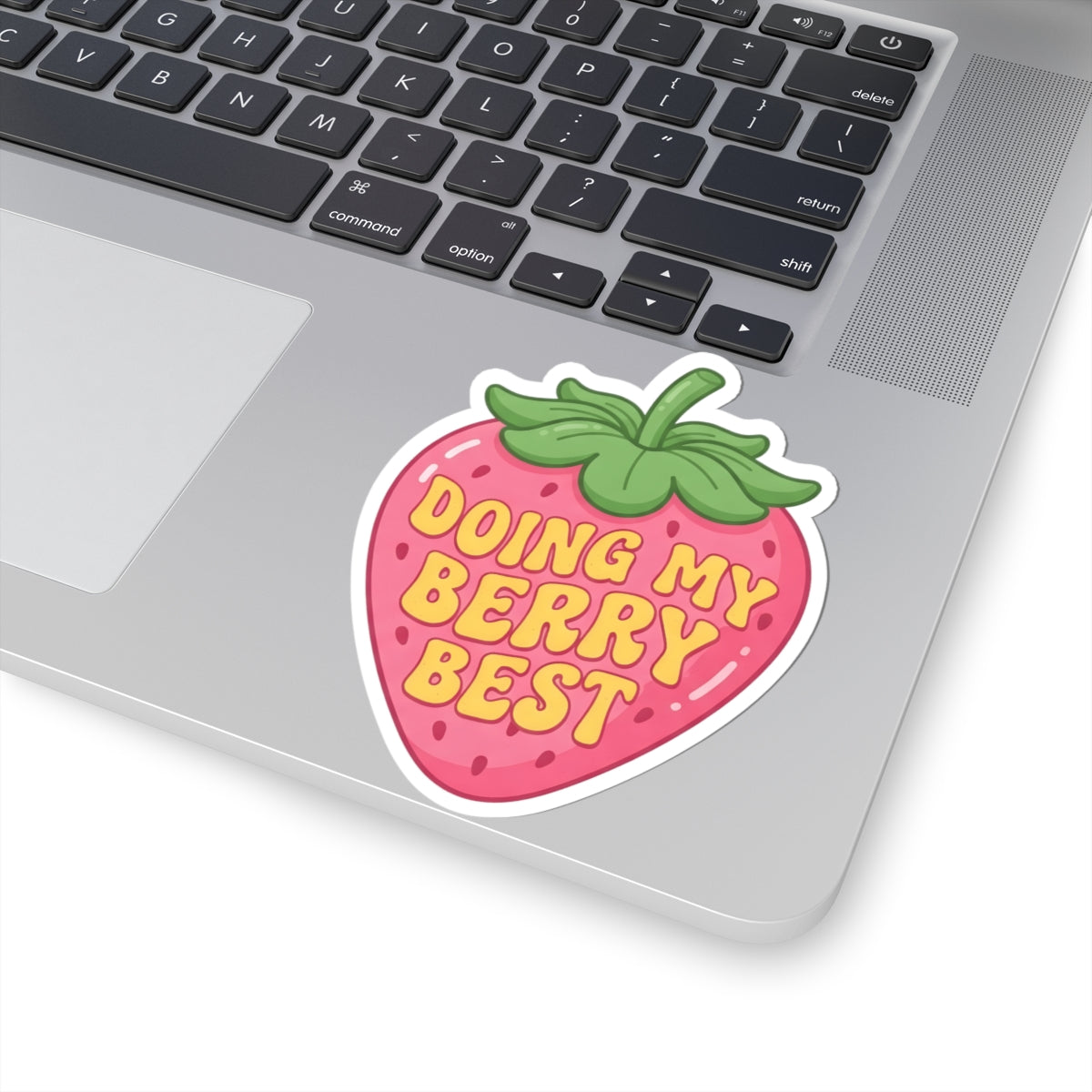 A kiss-cut strawberry sticker with the text 'DOING MY BERRY BEST' in yellow, placed on the palm rest of a silver laptop. The sticker has a white border and green leaves on top.